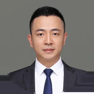 Professional headshot of an Asian man in a dark blazer, white dress shirt, and dark blue tie against a gray background